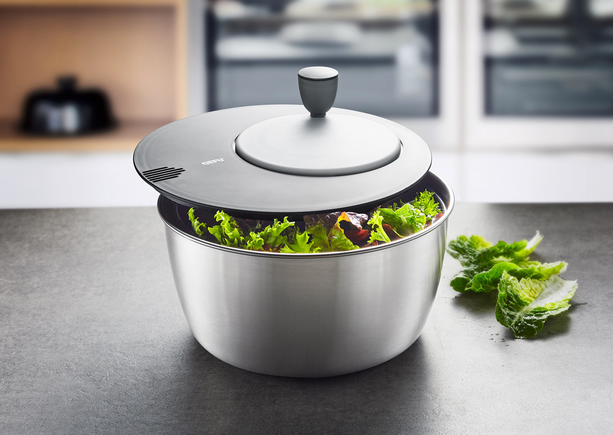 Salatschleuder mit Salat. Stahlschüssel und Deckel, grauer Griff. Küchenarbeitsplatte im Hintergrund.