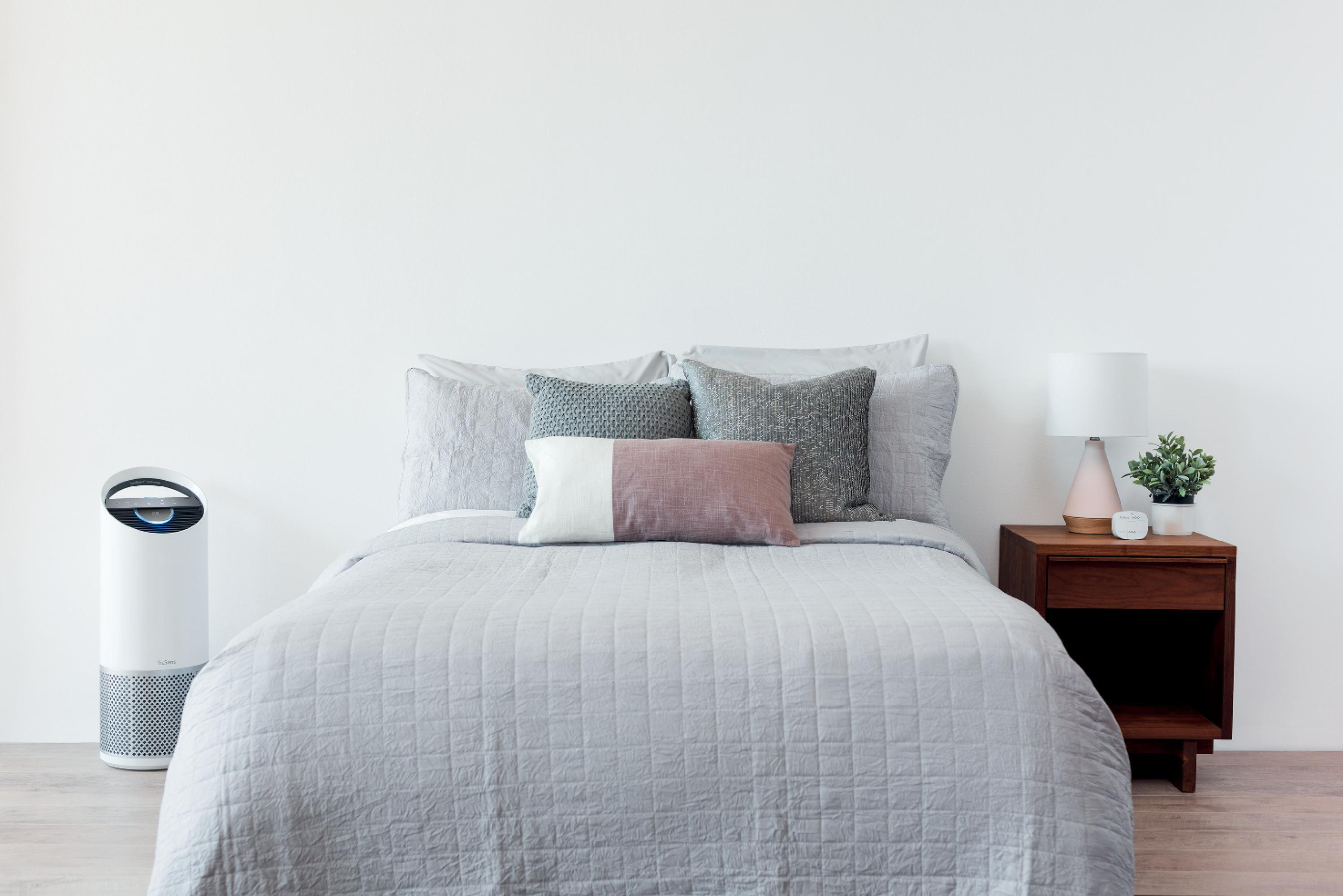 Ein Bett mit grauer Bettwäsche, Kissen, einem Beistelltisch mit Lampe und einem Luftreiniger links.