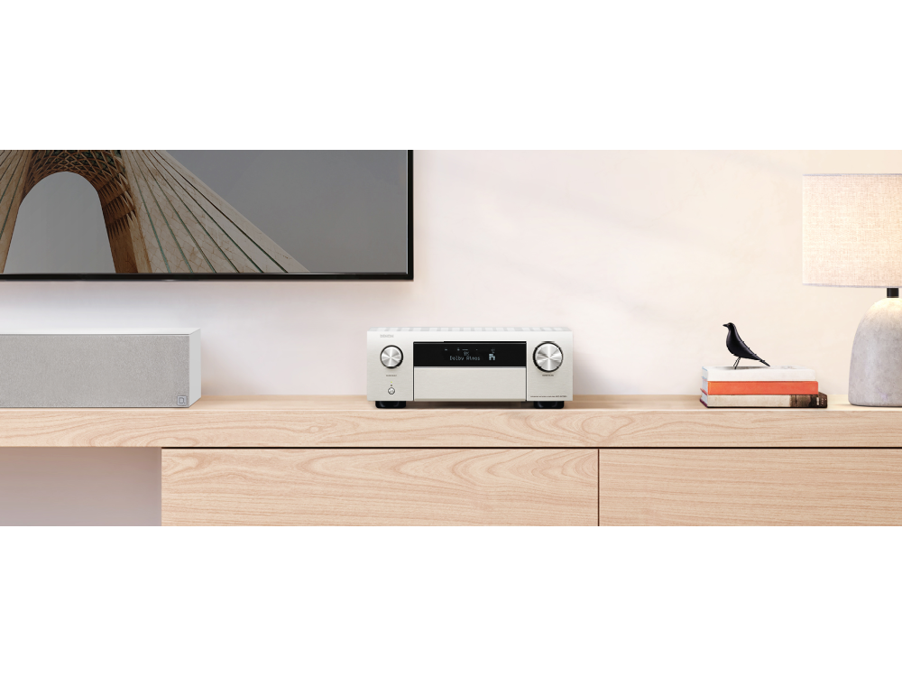 Receptor de audio Denon plateado en un mueble de madera. TV y altavoz en el fondo.
