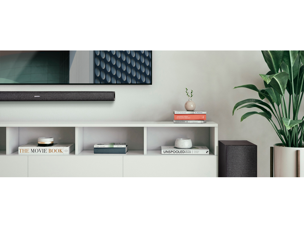 Salón con barra de sonido sobre un mueble blanco, una planta y un subwoofer.