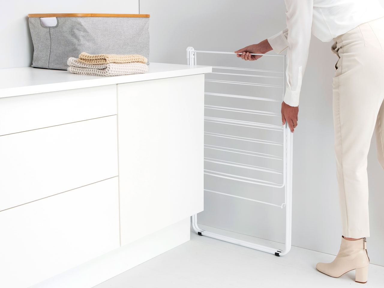 Une femme ouvre un étendoir blanc à côté d'un meuble blanc et d'un panier gris.