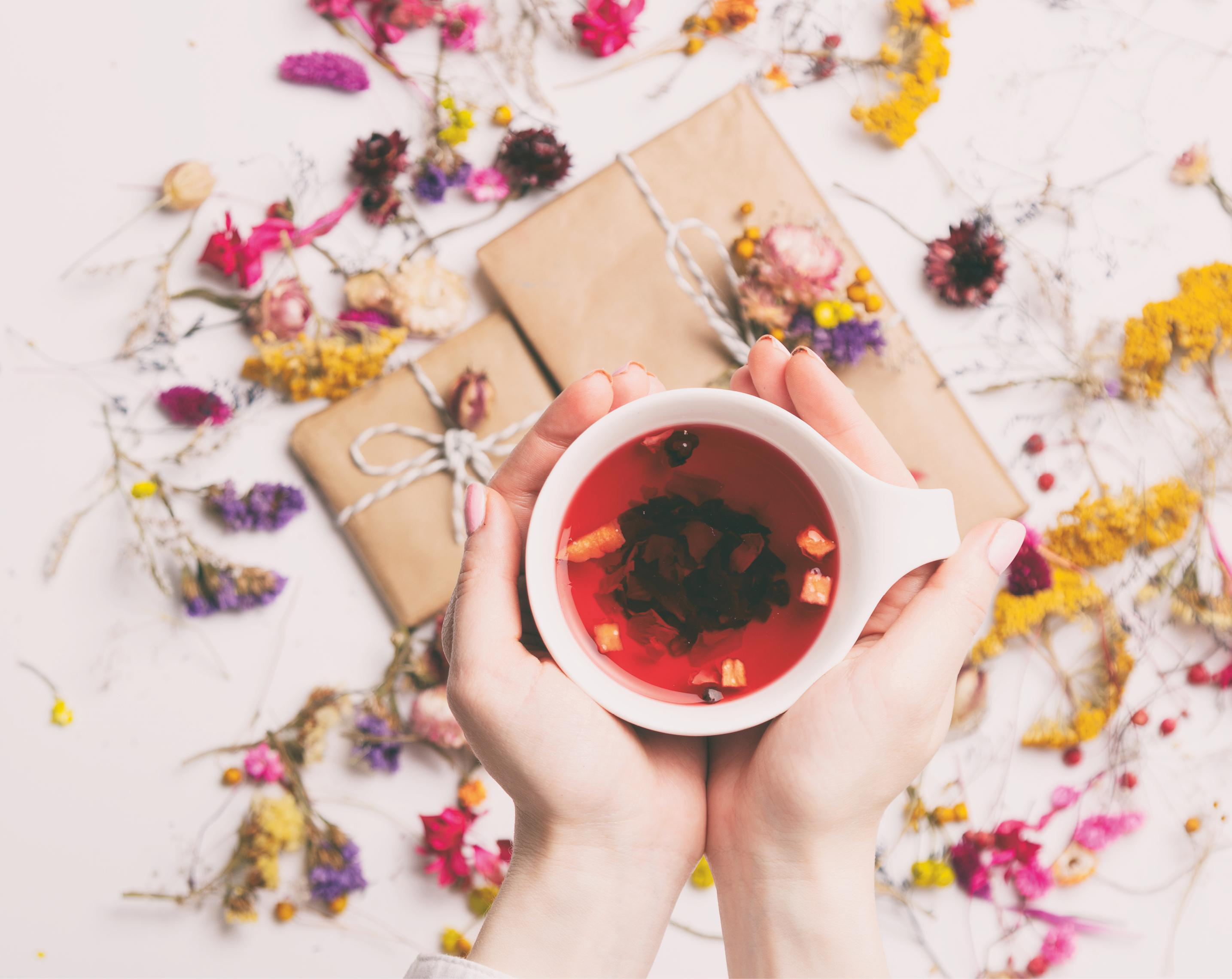 Hände, die eine weiße Tasse Tee halten, mit Blumen und verpackten Geschenken drumherum.