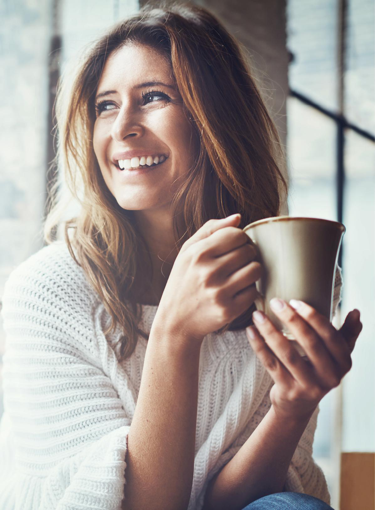 Eine lächelnde Frau, die eine Tasse Kaffee an einem Fenster hält.
