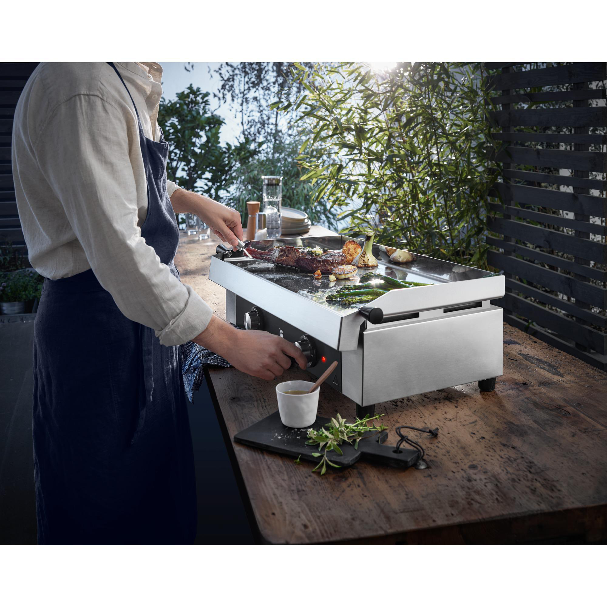 Eine Person grillt Essen auf einem Edelstahl-Außengrill auf einem Holztisch.