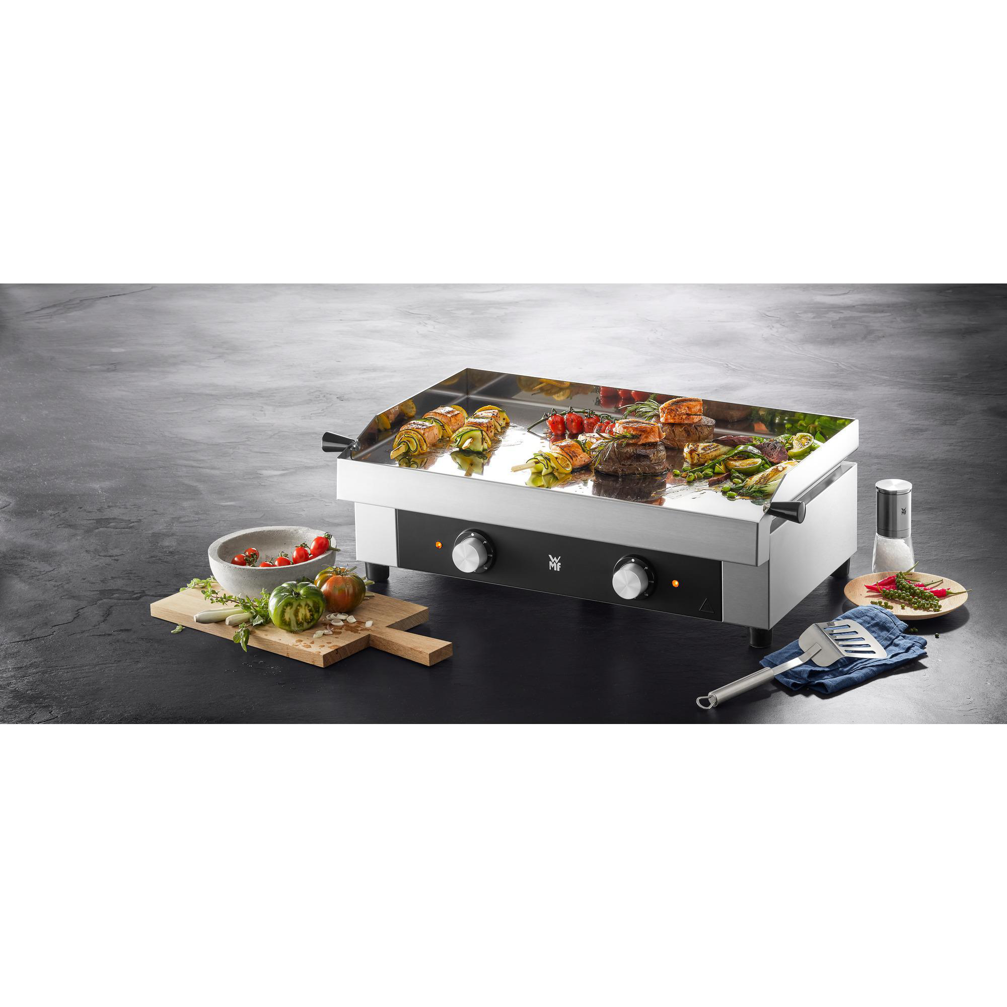Ein Elektrogrill mit gekochten Speisen. Einige rohe Gemüse liegen auf einem Holzbrett.