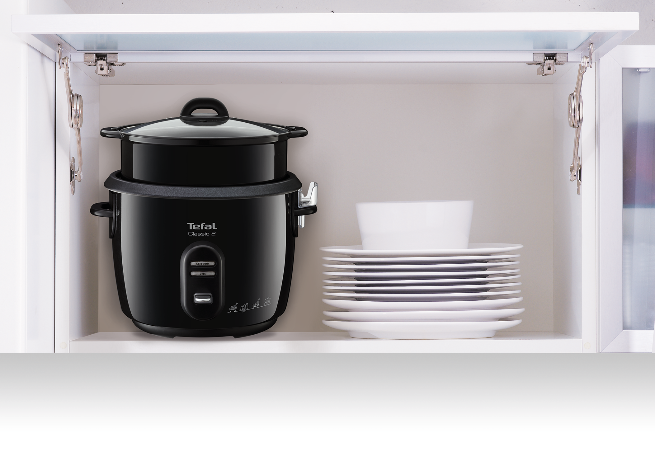 Black rice cooker with a glass lid. It's placed in a white cabinet with plates.
