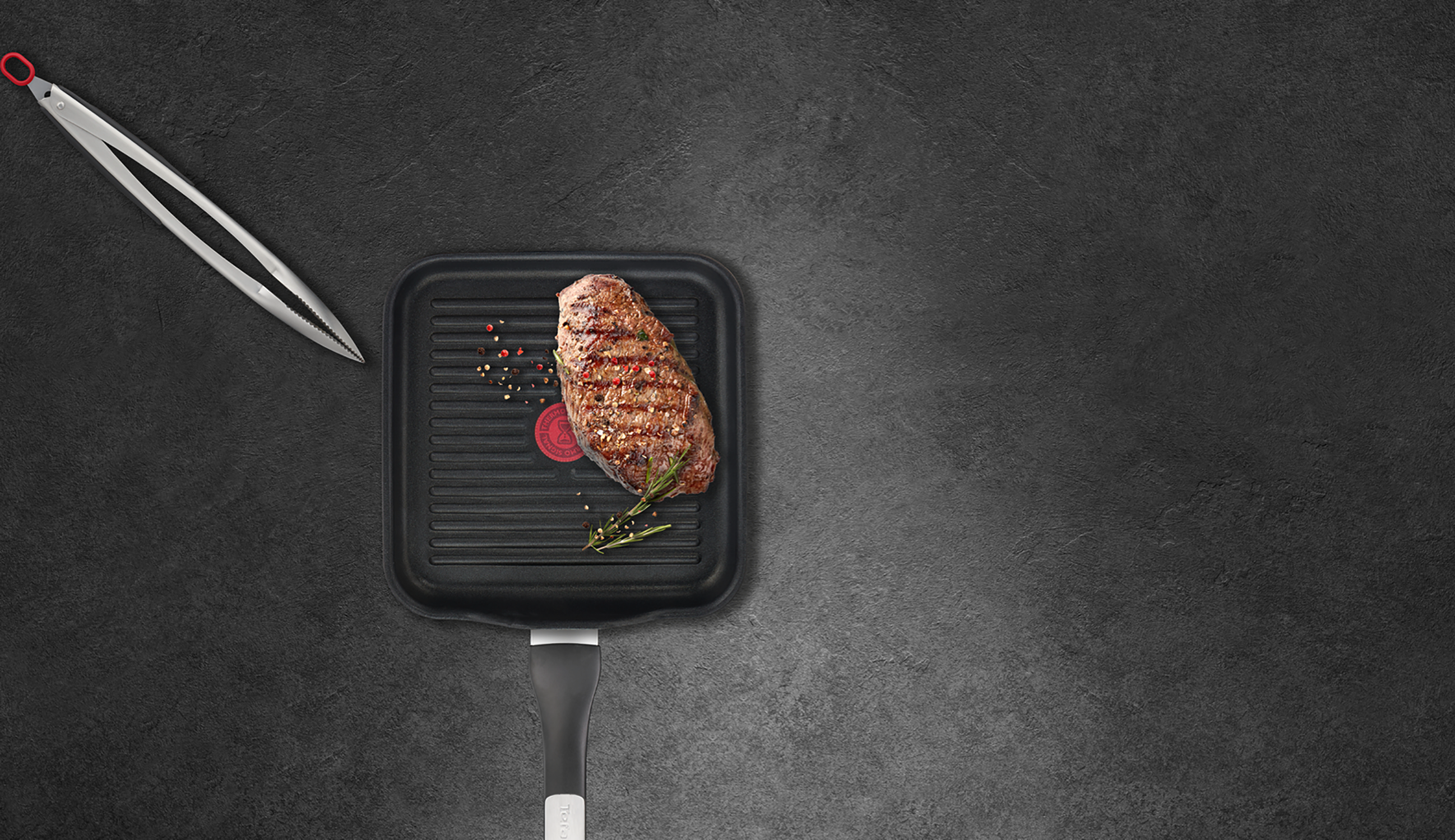 Steak on a square grill pan with tongs and spices, on a dark surface.