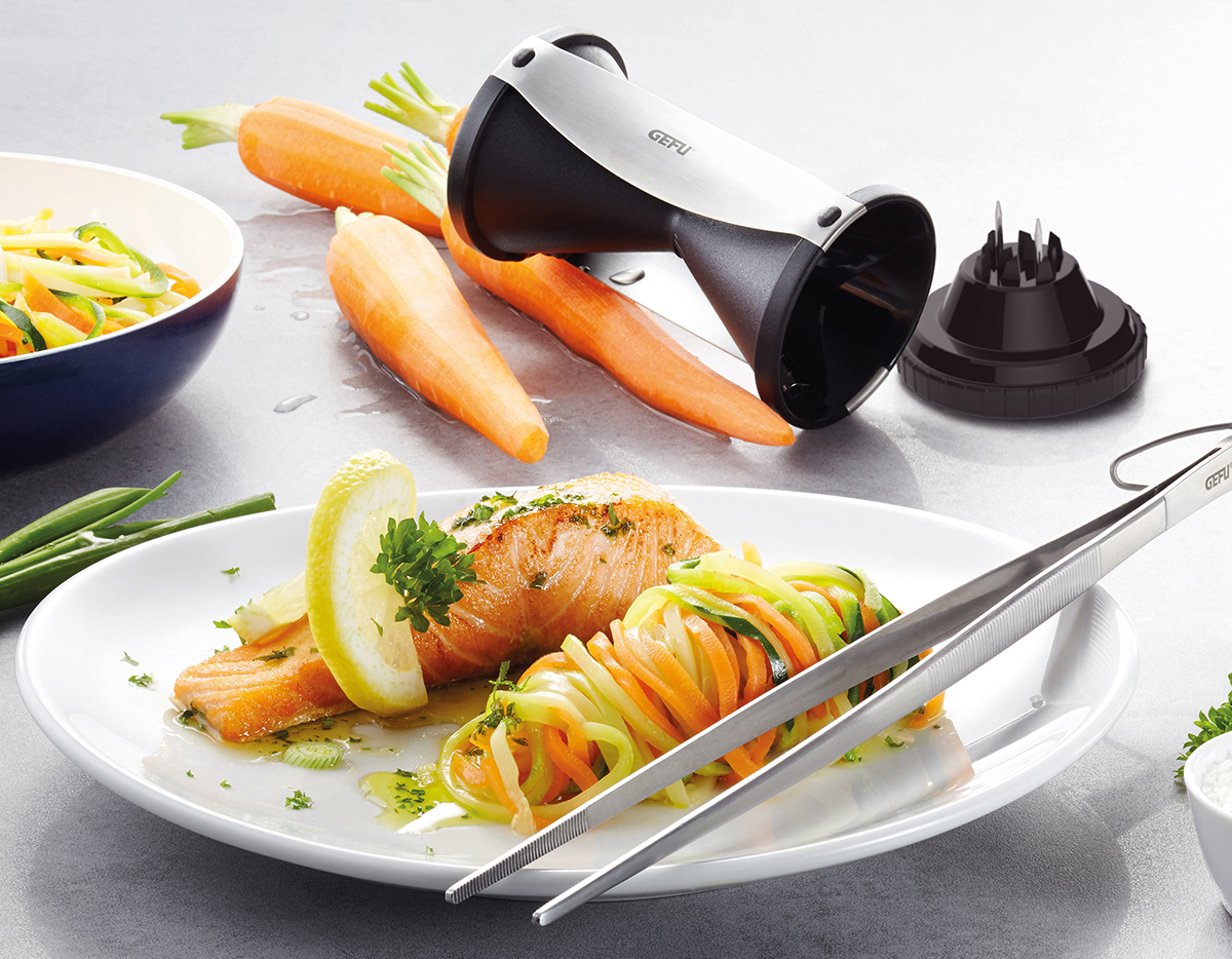 Ein Teller mit Lachs, Zucchini-Nudeln, Karotten und Zitrone. Kochutensilien befinden sich in der Nähe.