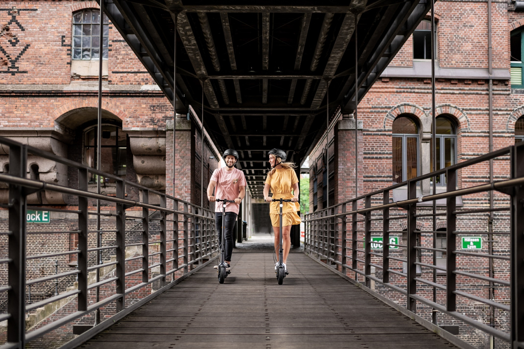 Zwei Personen auf Rollern auf einer Brücke. Der Hintergrund ist ein Backsteingebäude und eine Metallkonstruktion.
