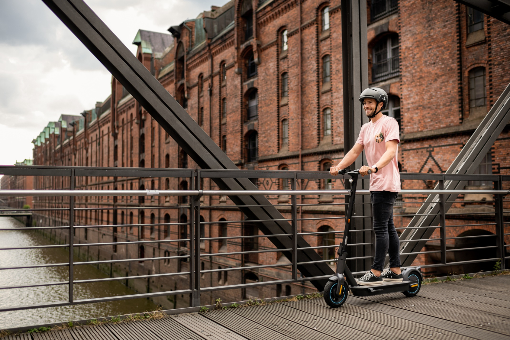 Mann auf einer Brücke, der einen Elektroroller fährt, mit einem Backsteingebäude im Hintergrund.