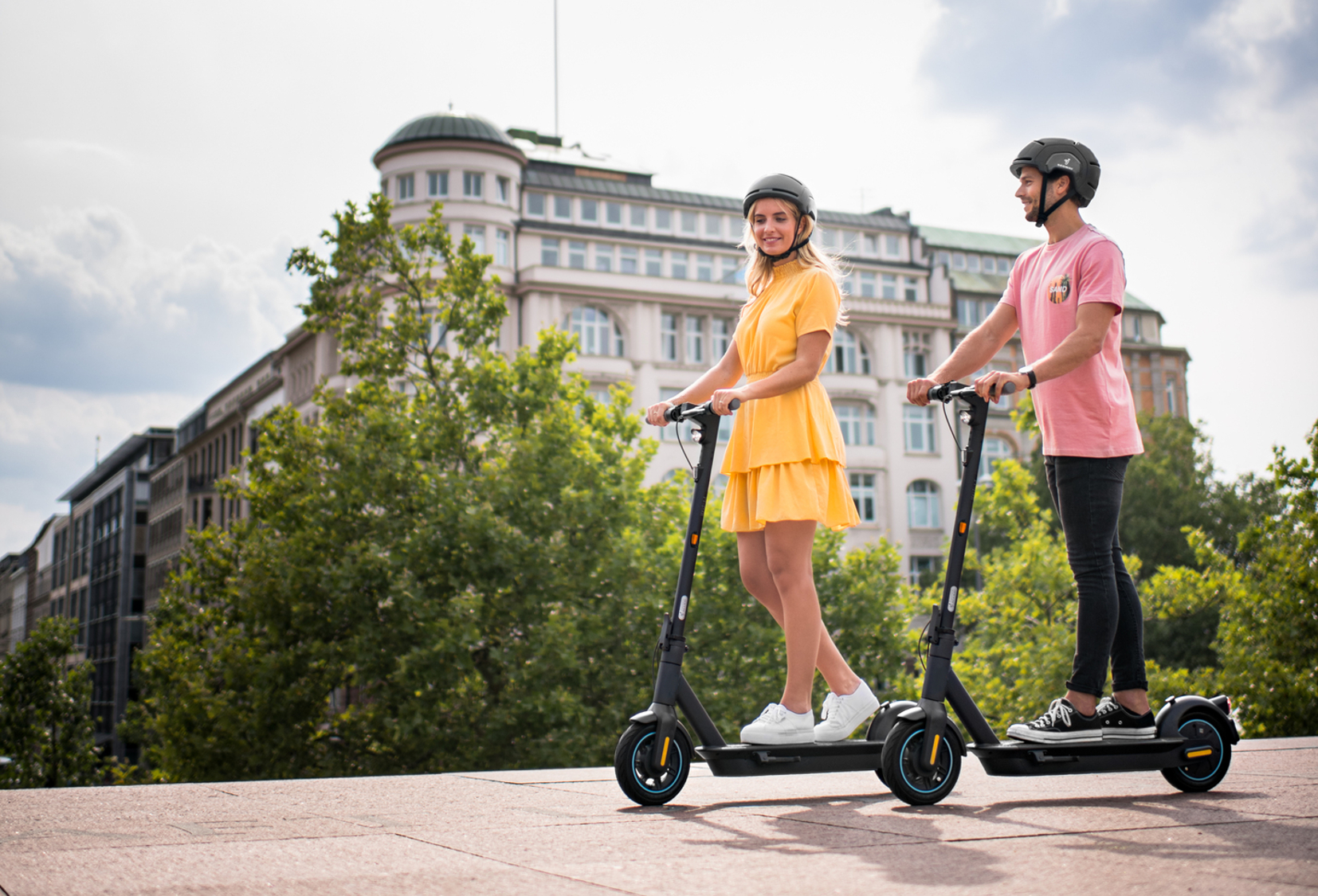 Zwei Personen fahren Roller auf einem Bürgersteig in der Stadt. Sie tragen Helme und im Hintergrund ist eine Stadt.