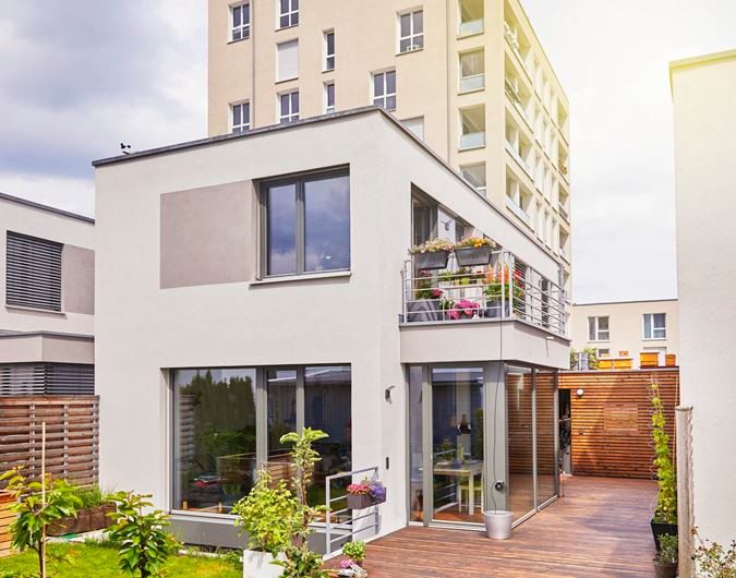 Modernes weißes Haus mit Balkon und Holzterrasse, neben einem hohen Apartmentgebäude.