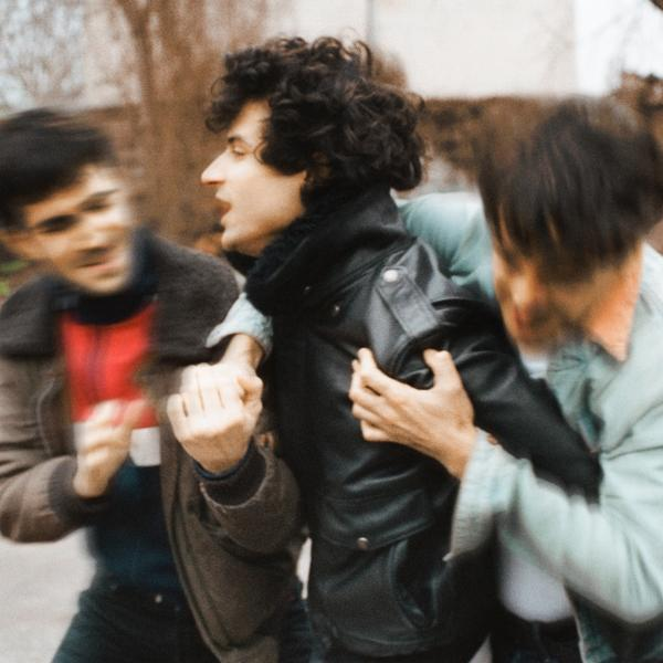 Three men are fighting. Two men restrain one, who is yelling. Blurry background of trees.