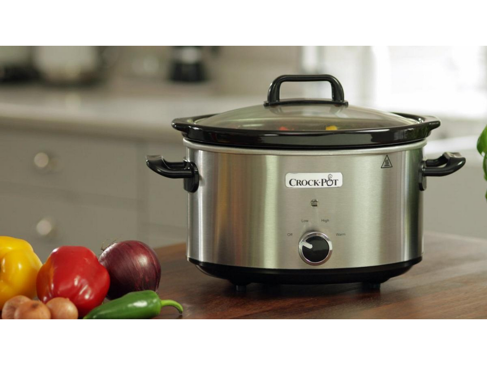 Olla de cocción lenta plateada con tapa de vidrio sobre una mesa de madera. Verduras en primer plano.