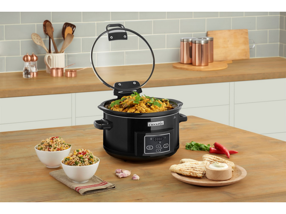 Una Crock-Pot negra con comida, dos cuencos de arroz y algunos acompañamientos sobre una mesa de madera.