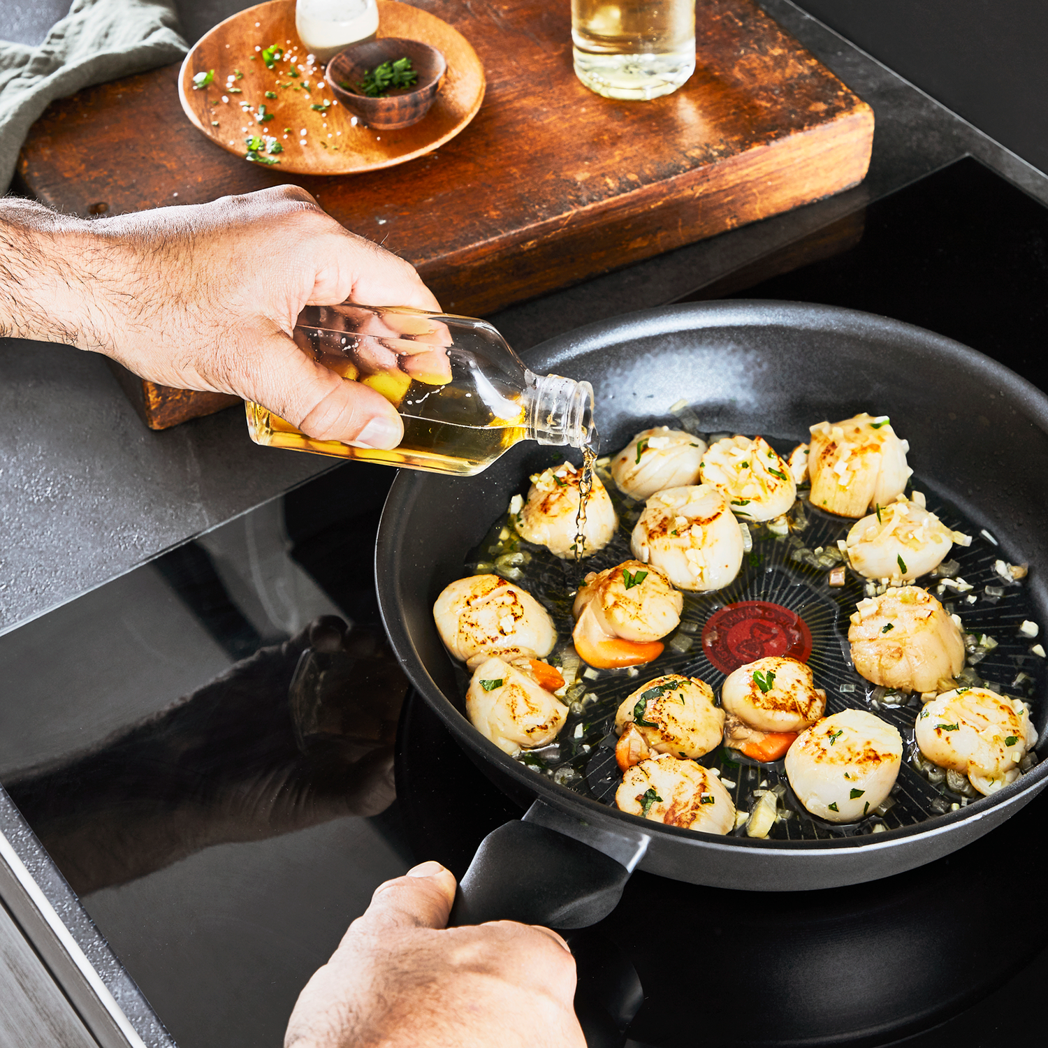 A person pouring oil into a pan with scallops. The pan is on a black stovetop. Other items are present.