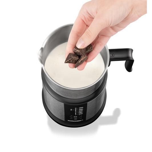 Eine Hand lässt Schokoladenstücke in einen Topf mit Milch fallen. Der Topf ist aus Edelstahl, der Hintergrund ist weiß.