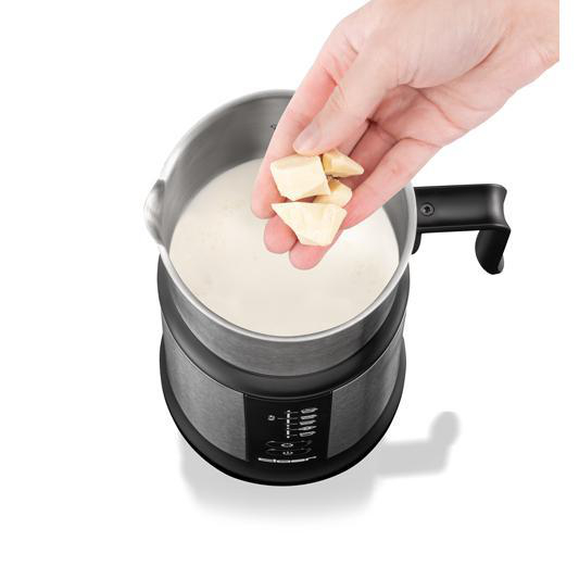 Hand fügt weiße Schokoladenstücke in einen silbernen Milchaufschäumer mit Milch ein.