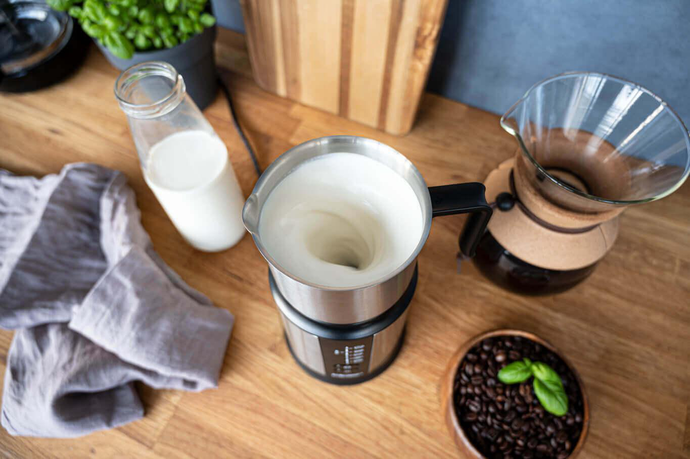 Milchaufschäumer mit Milch, eine Glasflasche Milch und Kaffeebohnen auf Holzoberfläche.