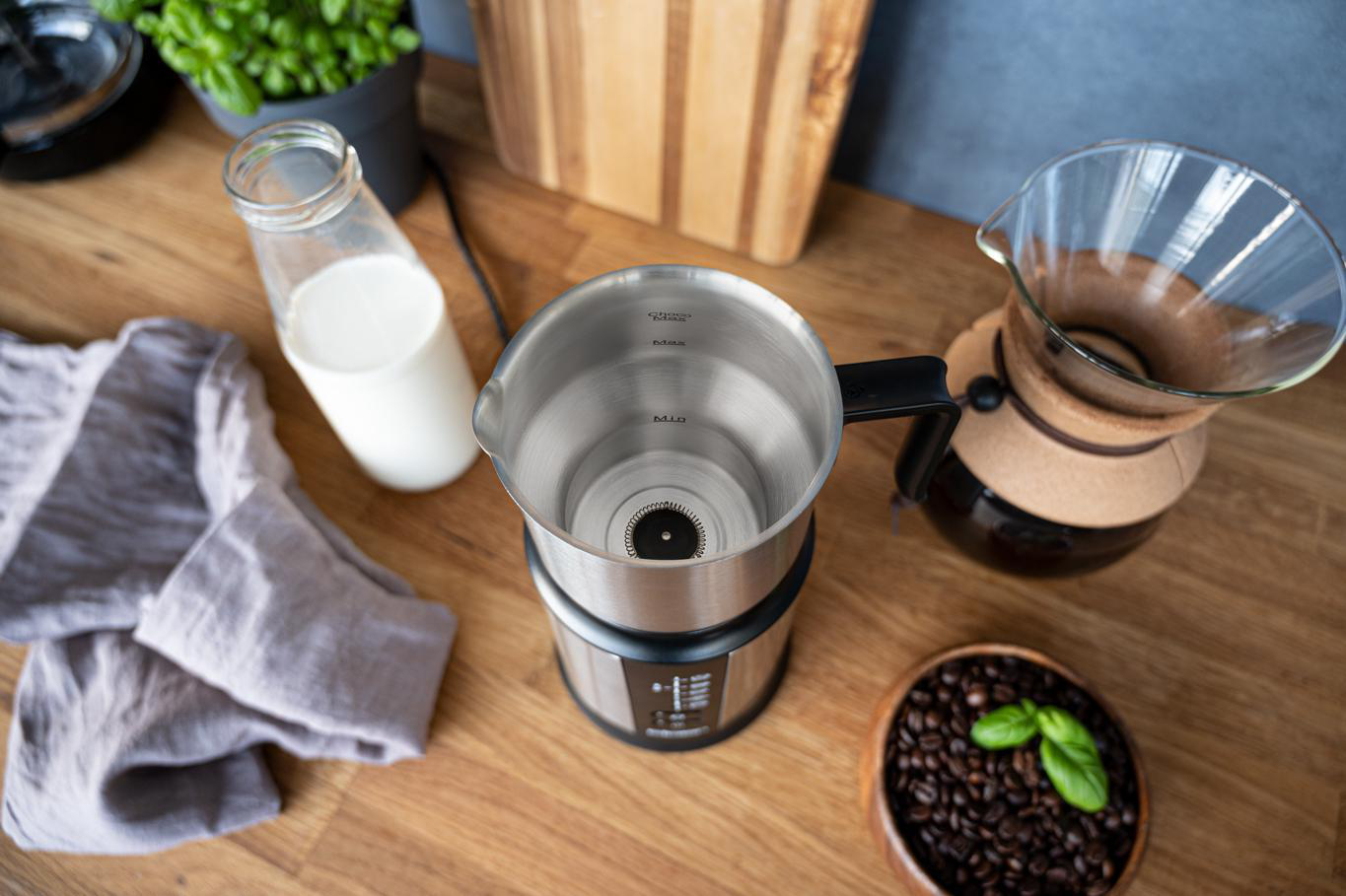 Ein Milchaufschäumer mit Milchflasche, Kaffeebohnen und eine Kaffeemaschine auf Holzoberfläche.