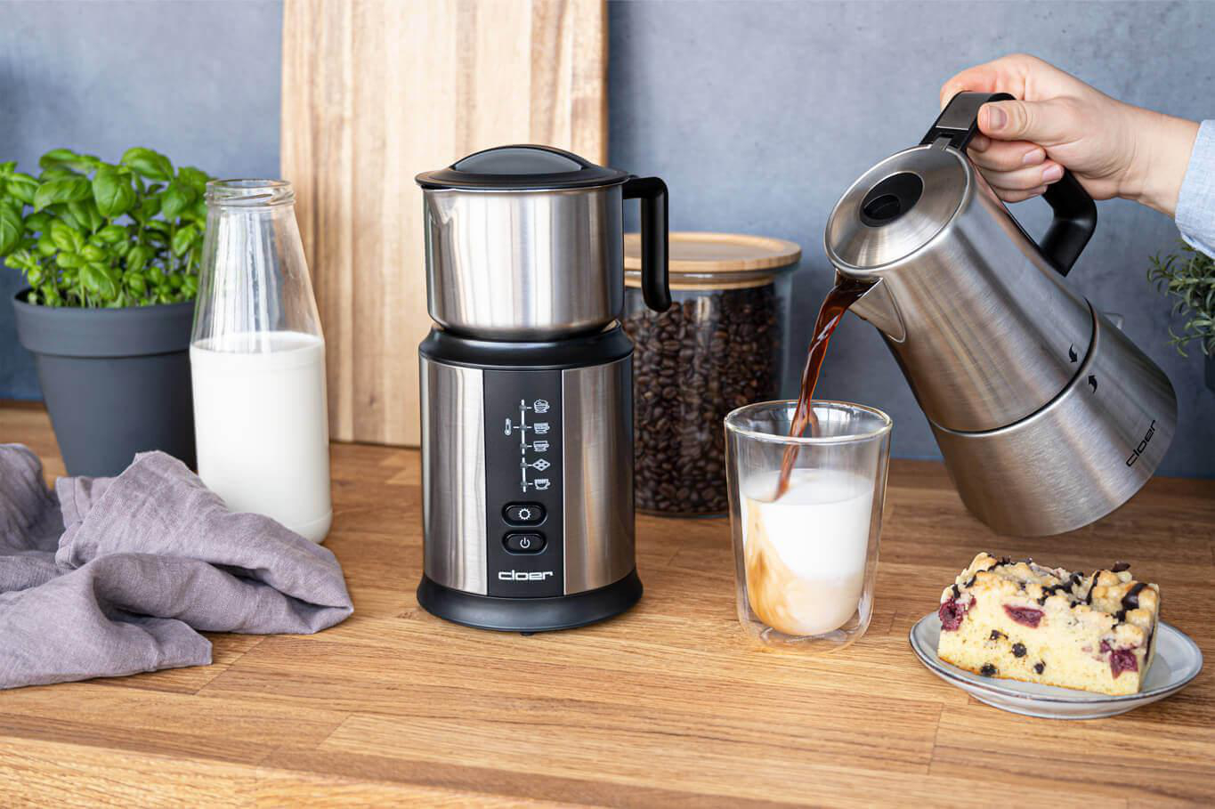 Eine Person gießt Kaffee aus einer Edelstahlkanne in ein Glas Milch mit Kaffeemaschine.