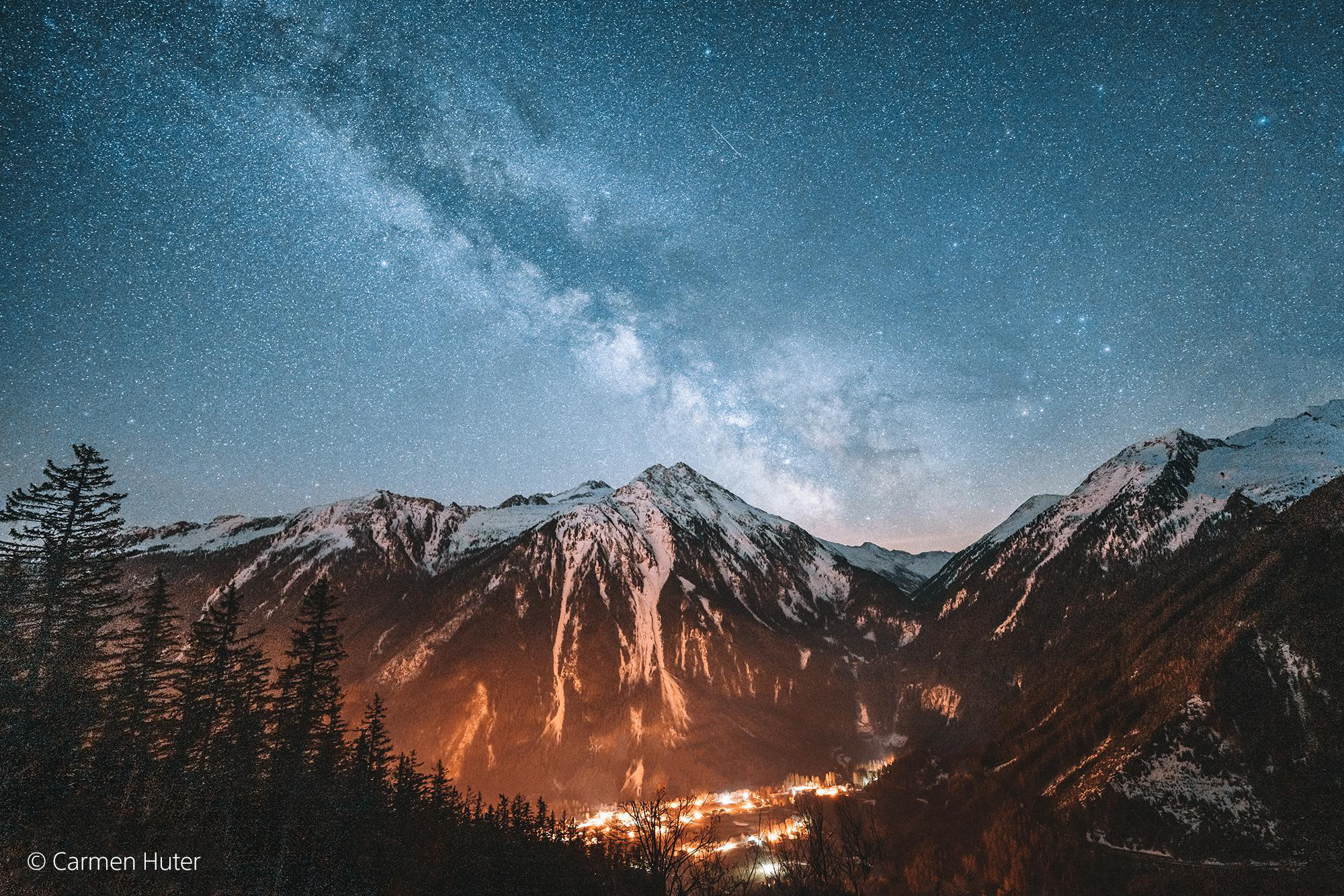 Nachtlandschaft: Bergkette, Sternenhimmel und Dorflichter am Fuße. Dunkler Vordergrund.
