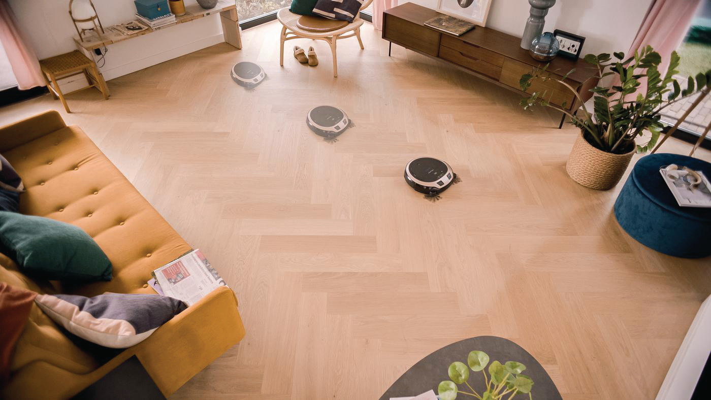 Drei Roboter-Staubsauger reinigen einen Holzboden. Gelbes Sofa und Pflanze.