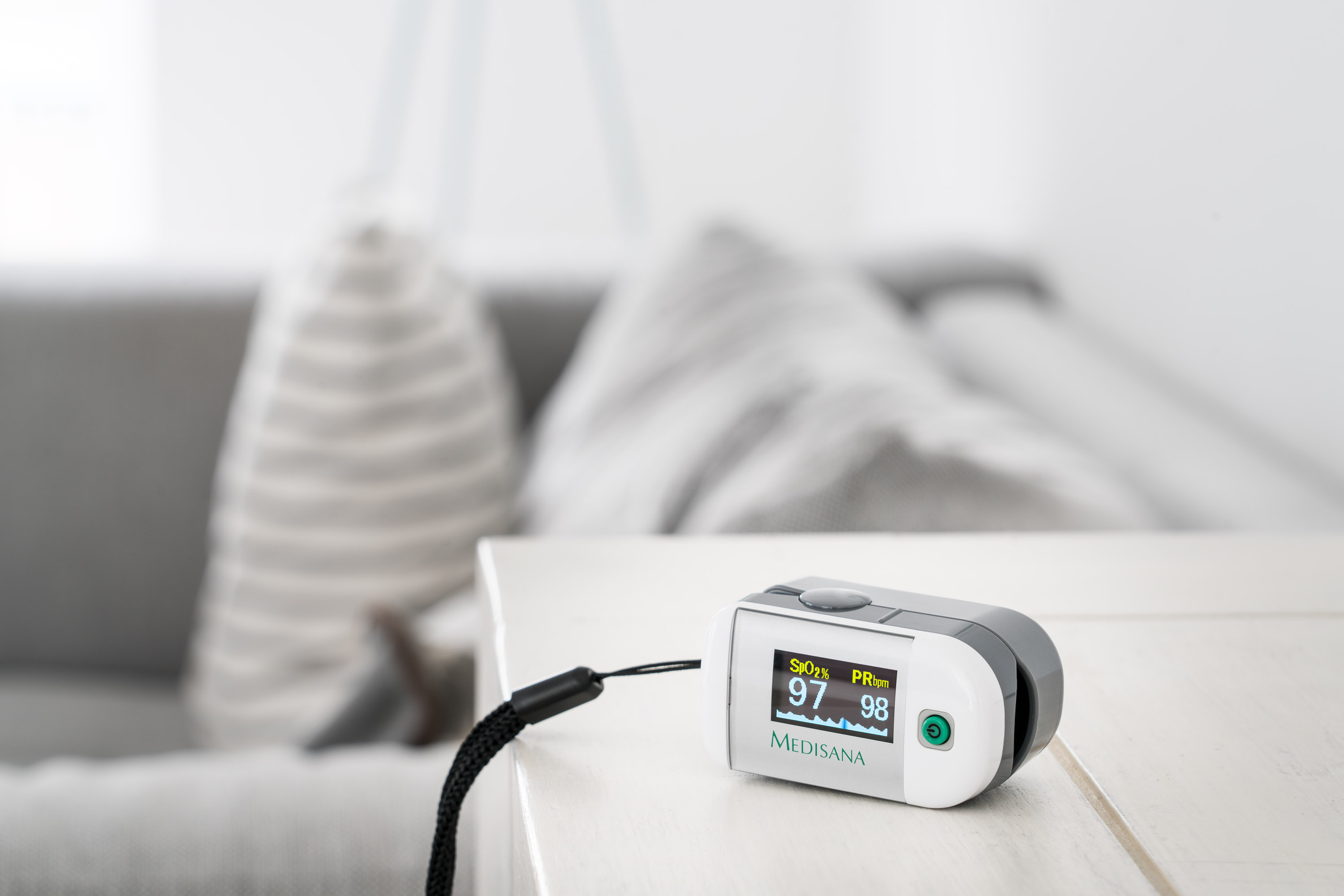 Pulsoximeter auf einem weißen Tisch. Anzeige: SpO2 97%, PR 98. Hintergrund ist ein Sofa.