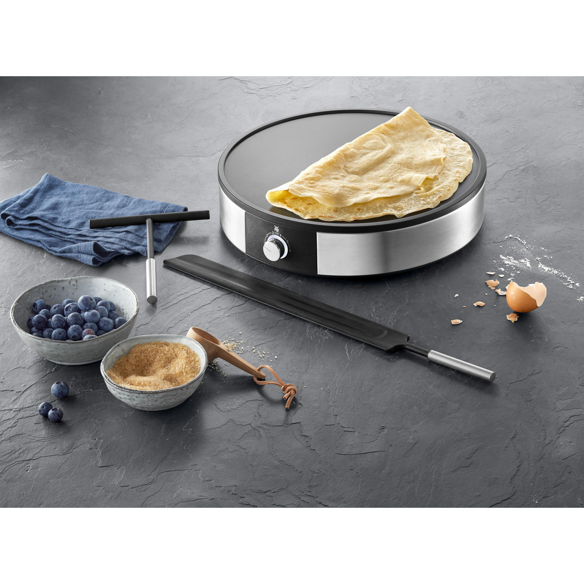 Ein Crêpes-Maker mit einem Crêpe, Werkzeugen und Zutaten auf einer grauen Oberfläche. Verschiedene Artikel sind um das Gerät herum platziert.