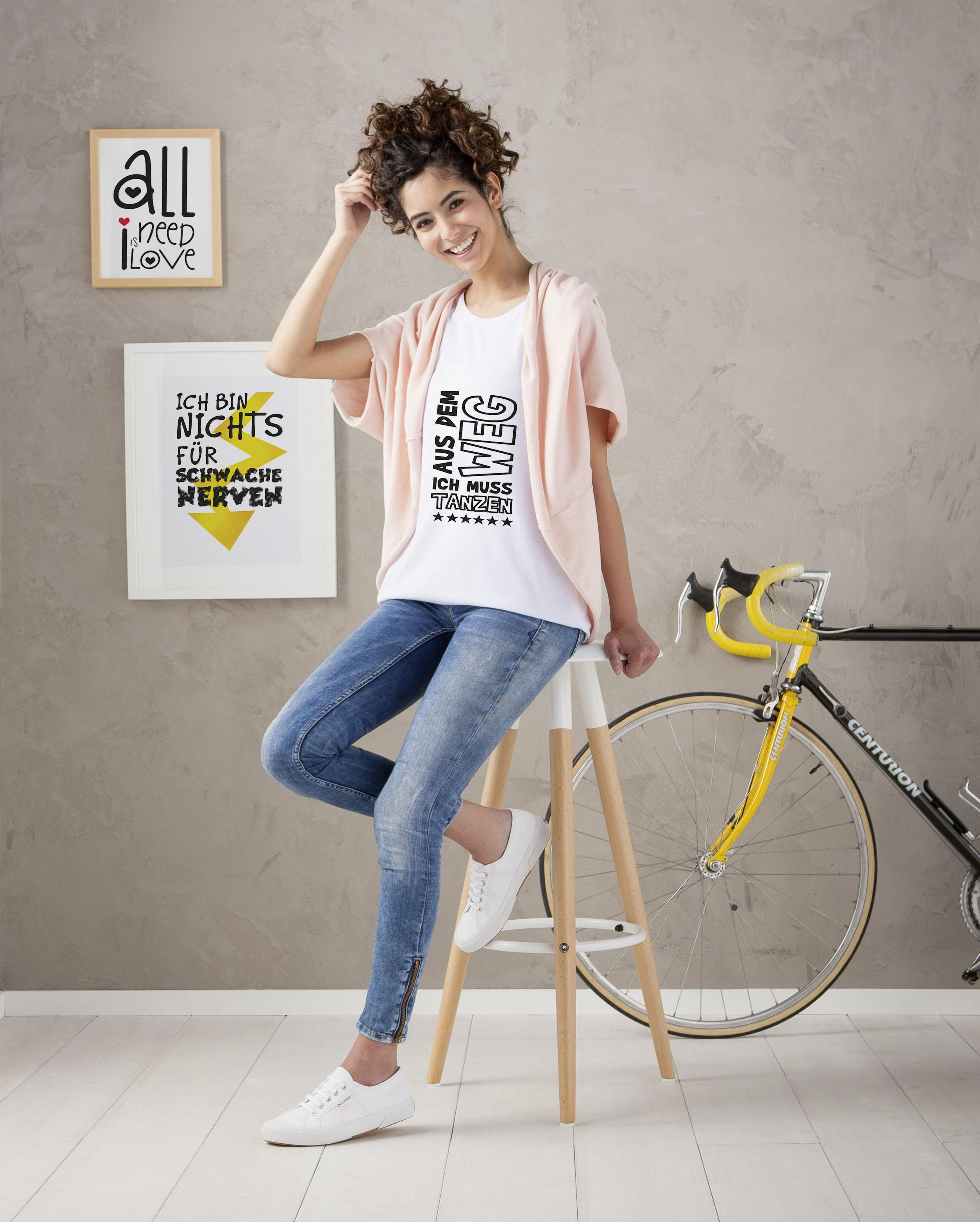 Eine Frau in weißem T-Shirt und Jeans, die sich auf einen Hocker lehnt. Kunstwerke und Fahrrad.