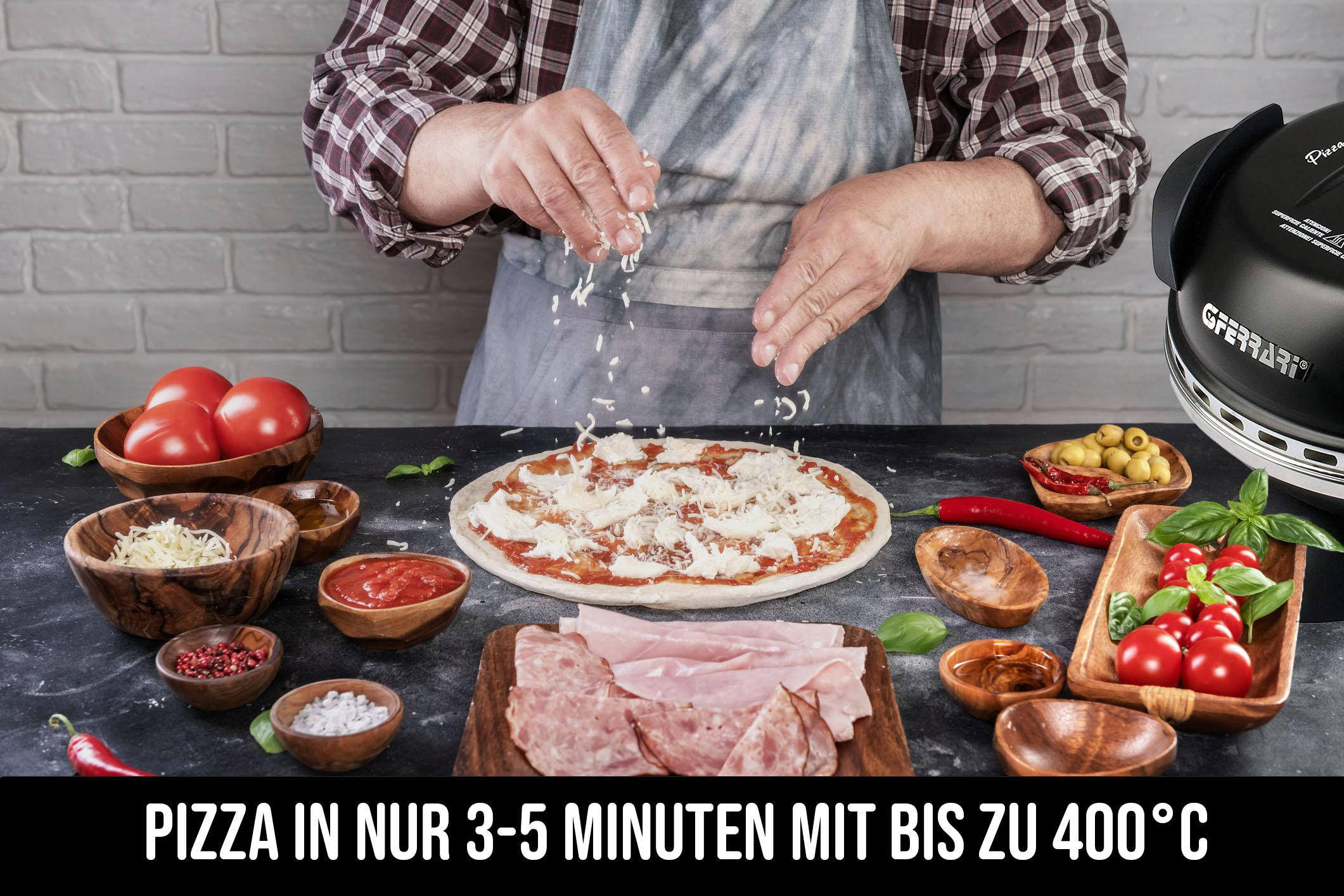 Eine Person bestreut einen Pizzaboden mit Käse, mit Tomaten, Schinken und einem Pizzaofen im Hintergrund.