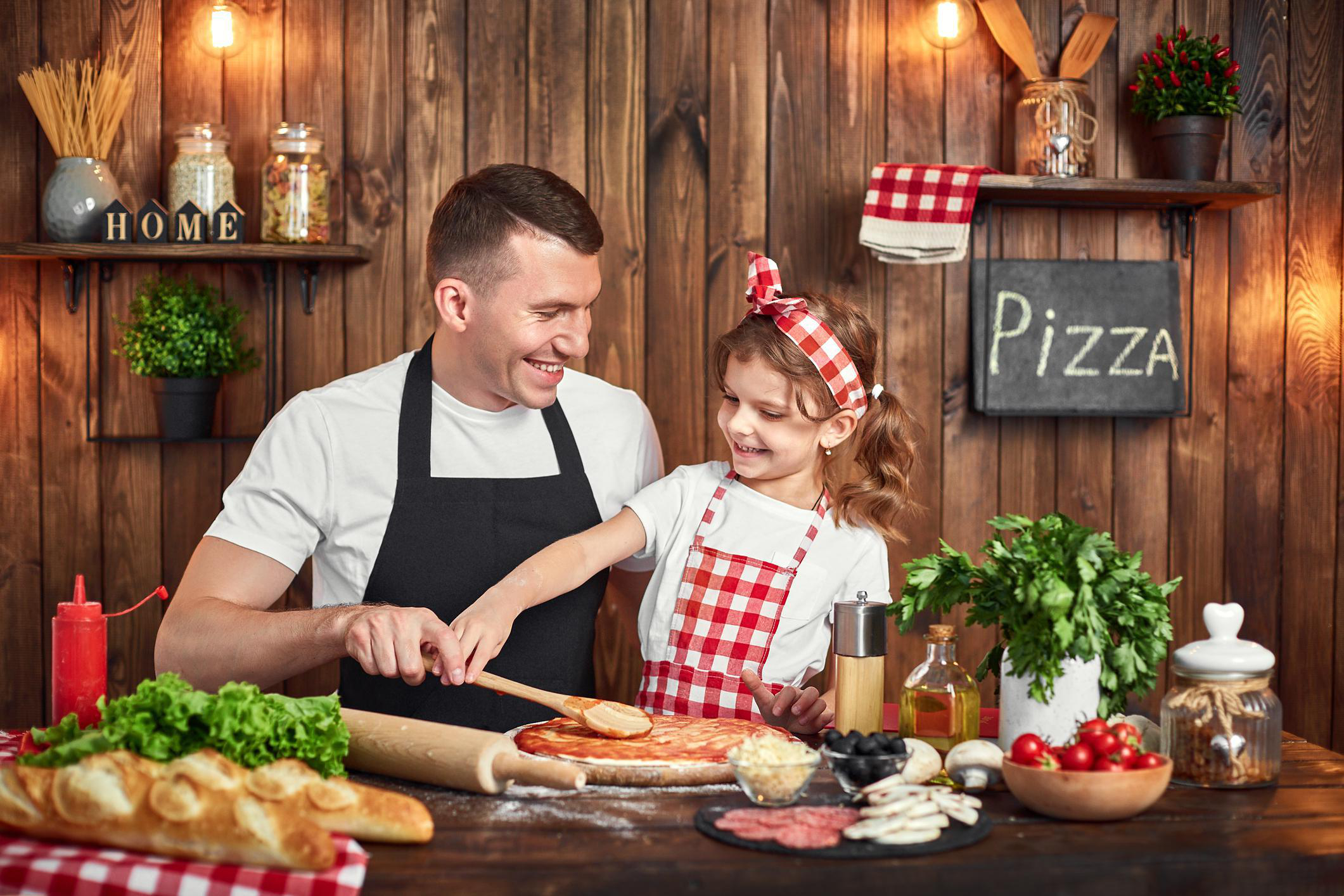 Ein Vater und seine Tochter machen Pizza. Der Vater lächelt, die Tochter verteilt Soße.