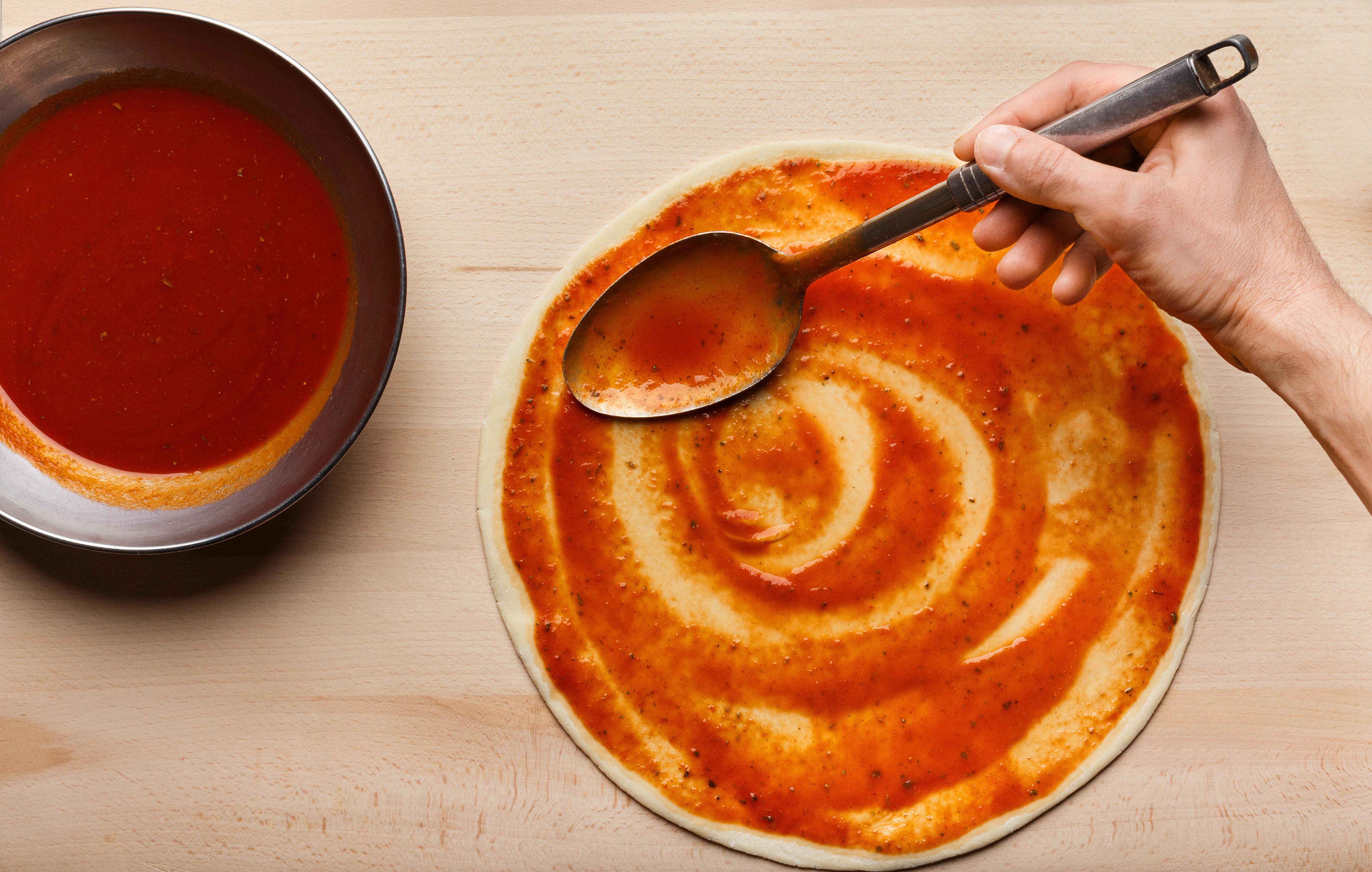 Die Hand einer Person, die mit einem Löffel Tomatensauce auf einen Pizzateig verteilt. Eine Pfanne mit Sauce ist daneben.