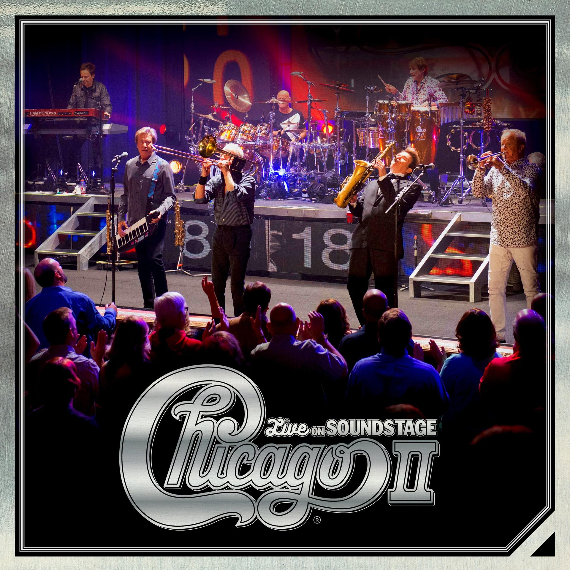 Live-Musikkonzert mit der Band Chicago. Bühne mit Musikern, Instrumenten und Publikum. Silbernes Logo unten.