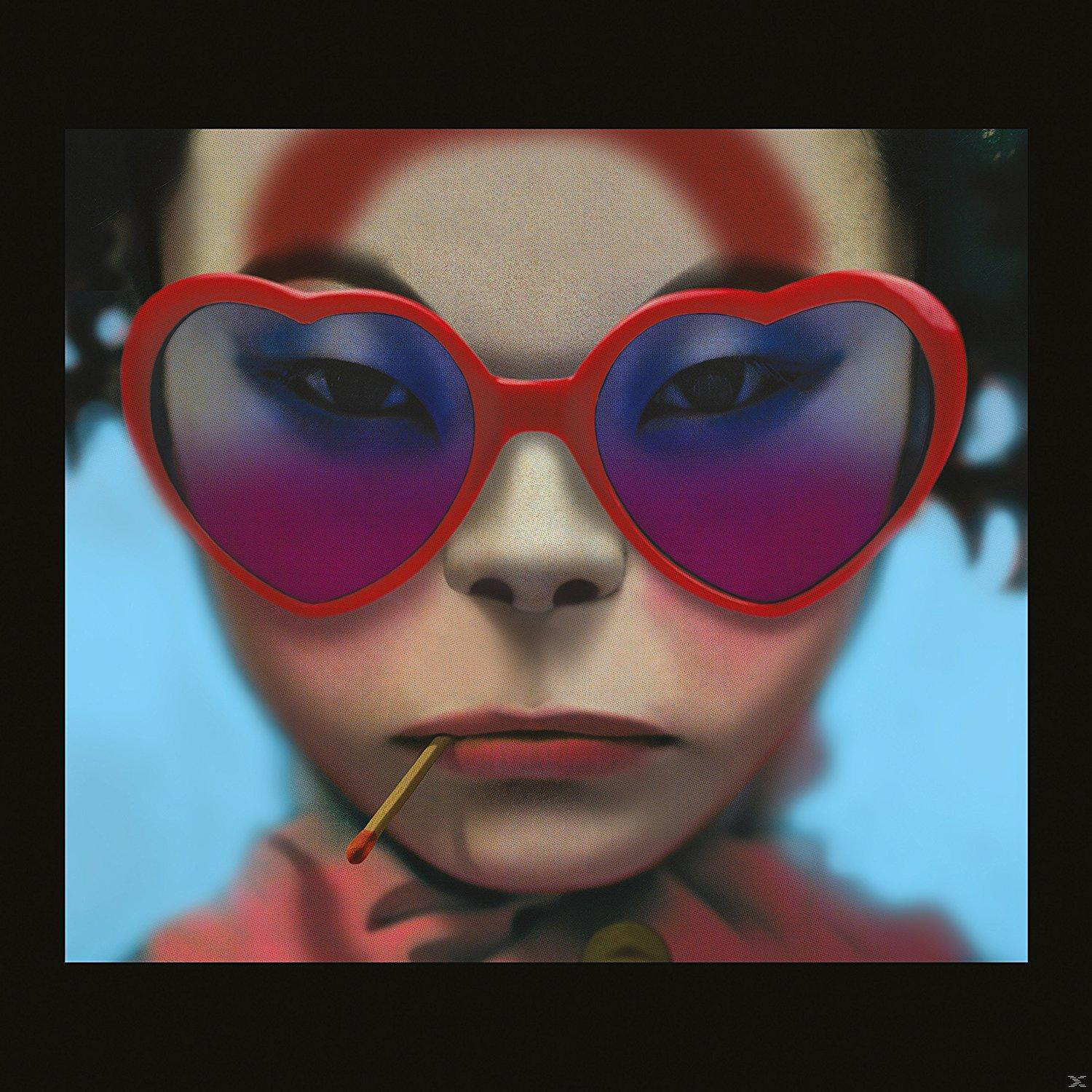 Nahaufnahme eines Gesichts mit roter herzförmiger Brille, einem Streichholz im Mund und einem Ziel auf der Stirn.