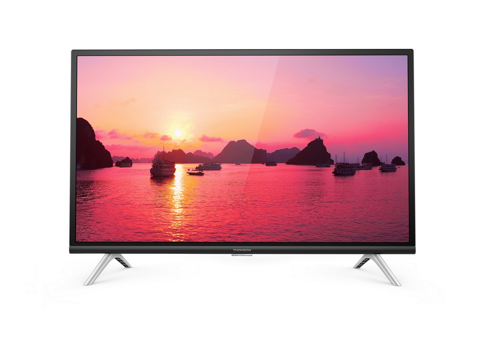 Un televisor de pantalla plana muestra una puesta de sol sobre el agua con barcos. El televisor tiene patas plateadas y un marco negro.
