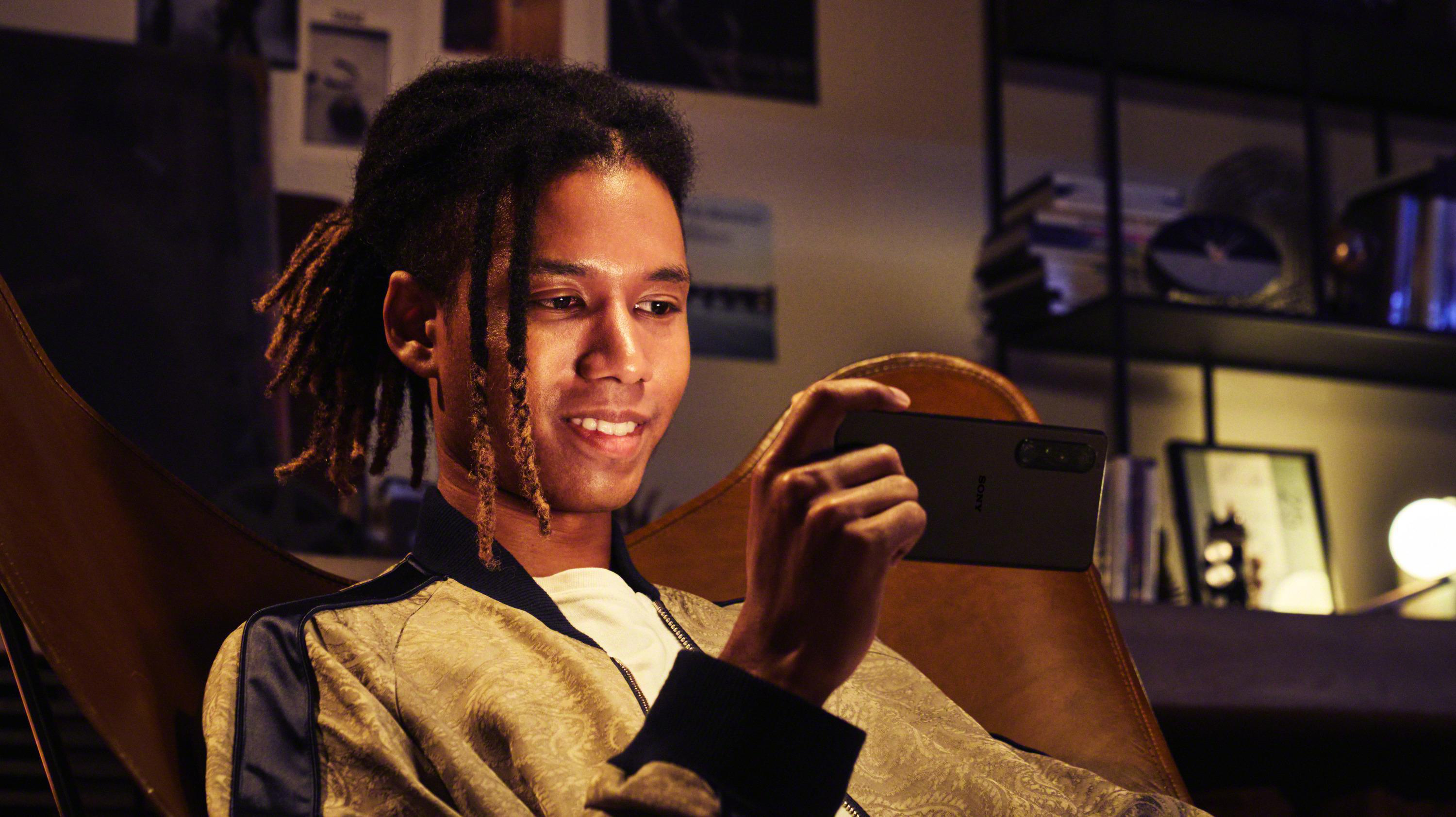 Ein junger Mann mit Dreadlocks lächelt, während er ein Telefon hält. Er sitzt in einem Stuhl, mit einem Regal im Hintergrund.