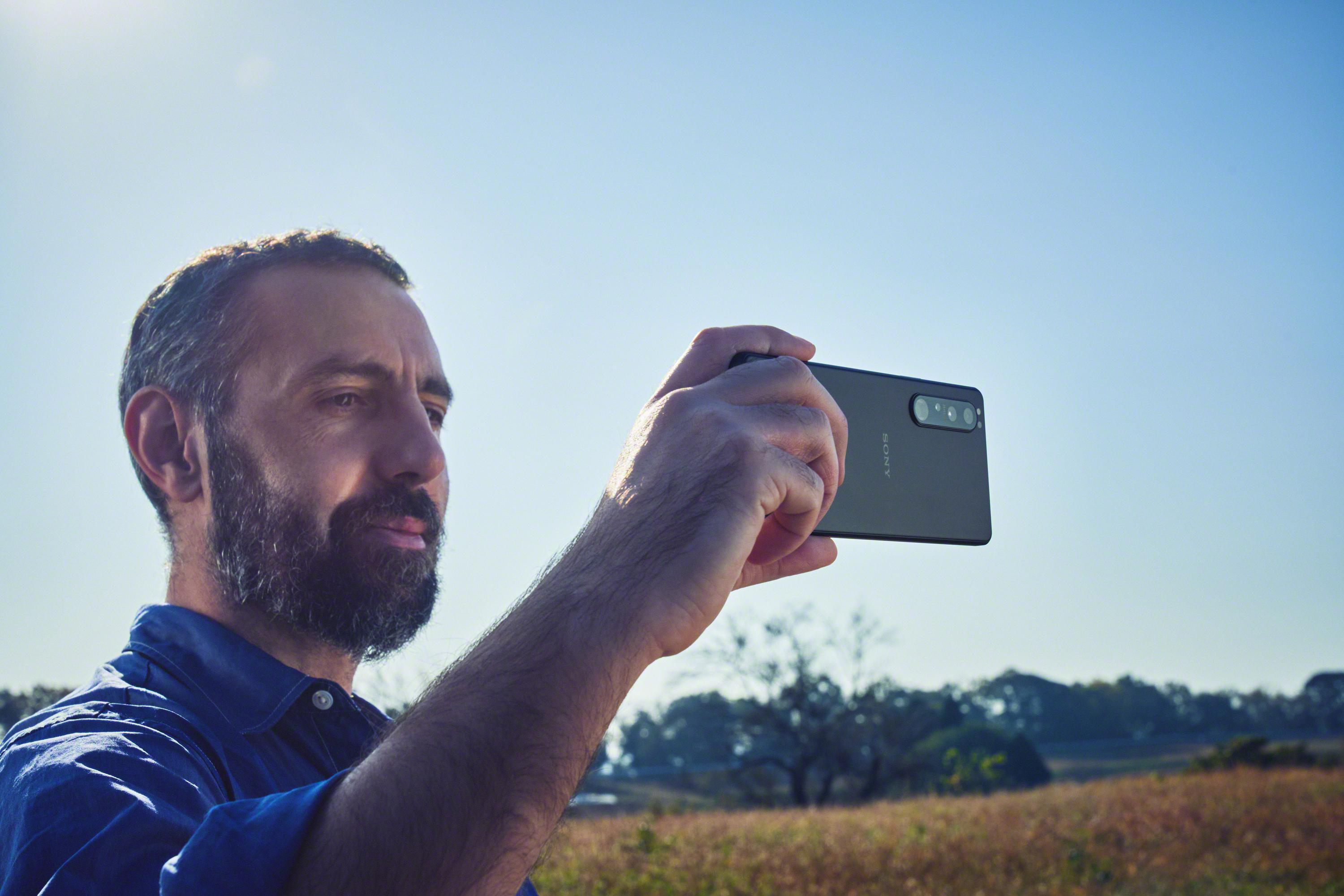 Ein Mann in einem blauen Hemd macht ein Foto mit einem schwarzen Telefon.