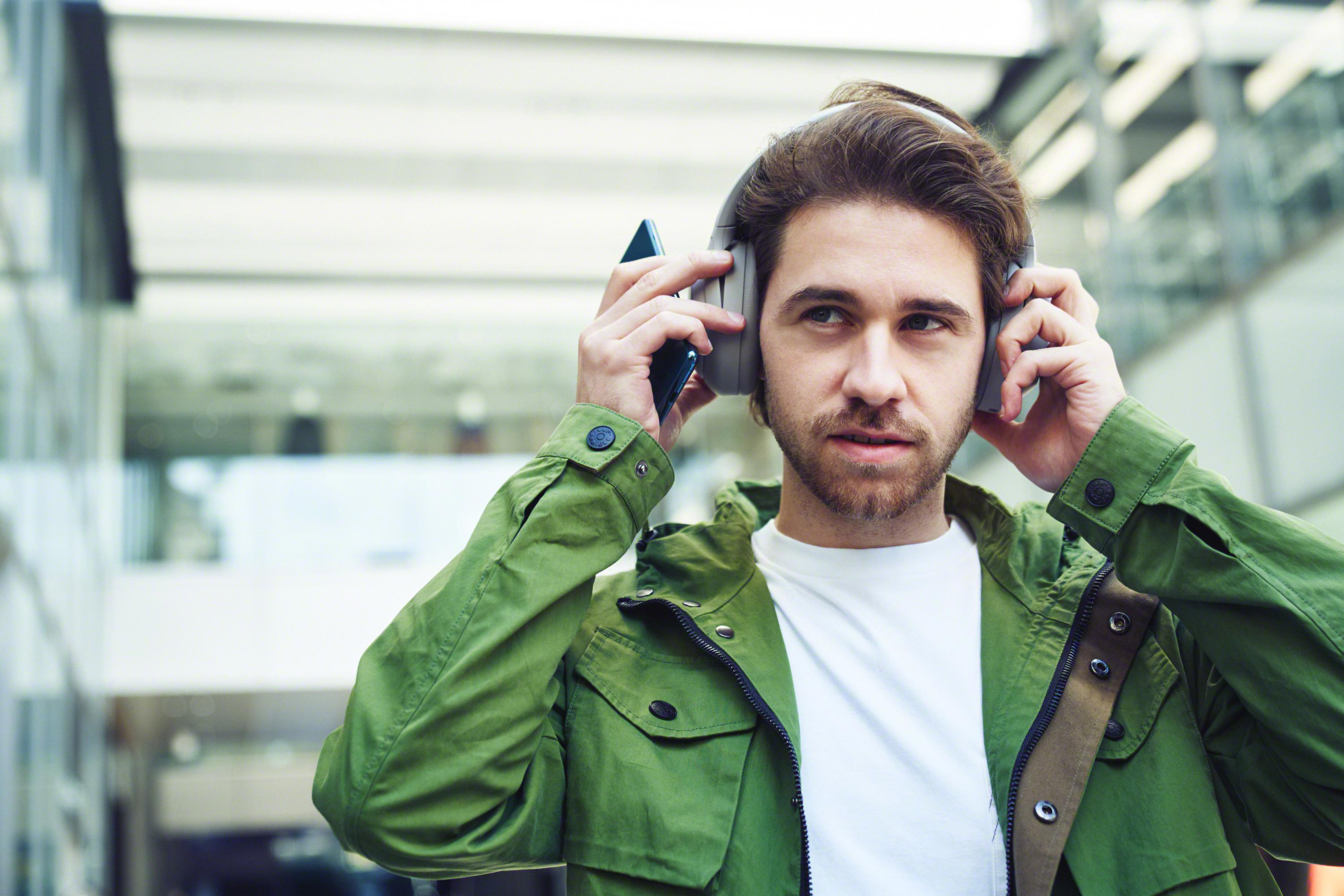 Ein Mann mit Bart setzt Kopfhörer auf, während er ein Smartphone hält. Er trägt eine grüne Jacke.