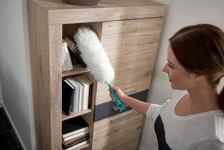 Eine Frau putzt einen Holzschrank mit einem grünen Staubwedel, Bücher im Inneren.
