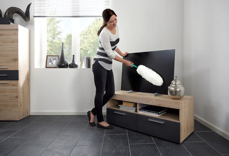 Eine Frau staubt einen Fernseher mit einem weißen Staubwedel ab. Der Fernseher steht auf einem Holzschrank.