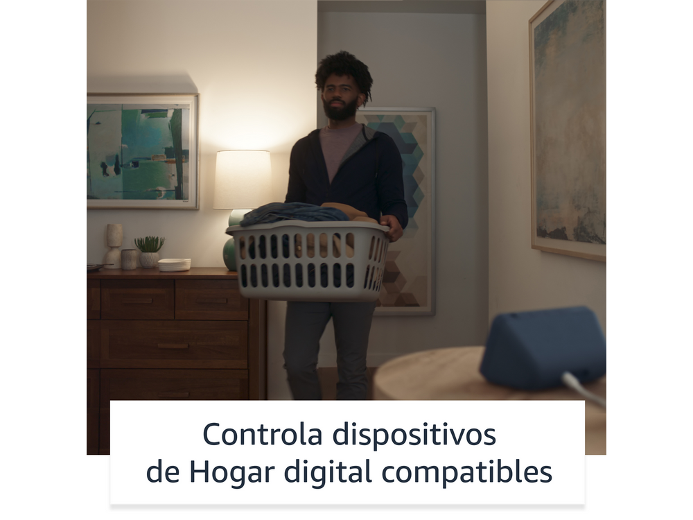 Hombre con cesta de ropa, de pie en una habitación. Altavoz inteligente en mesa. Controla dispositivos.