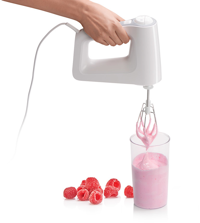 Een hand die een witte mixer vasthoudt en een roze mengsel in een glas met frambozen mengt.
