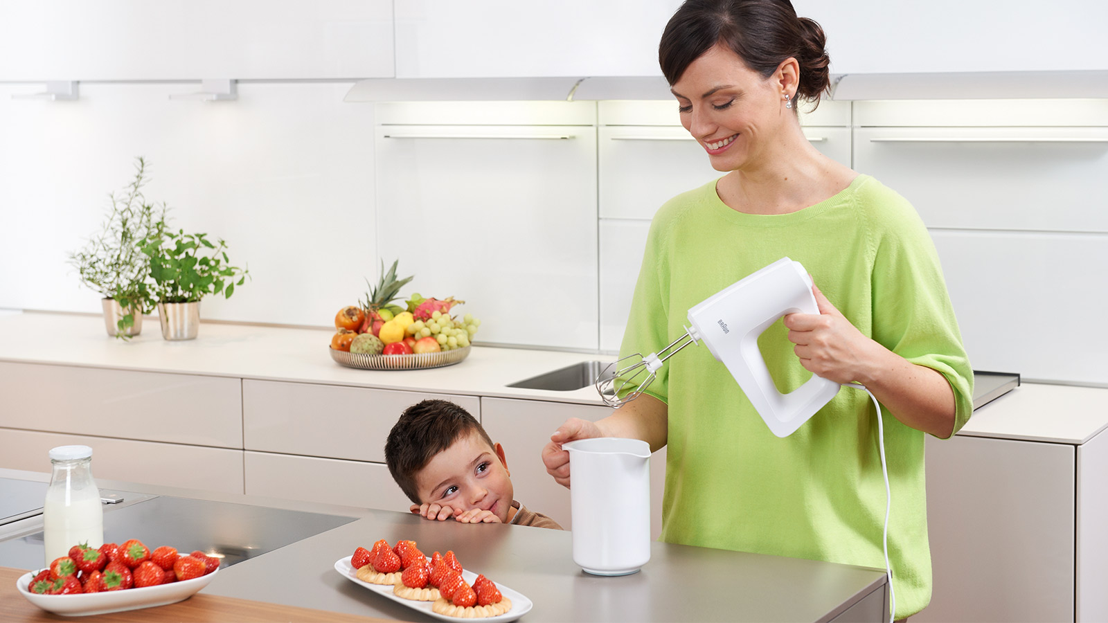 Een vrouw gebruikt een witte handmixer. Een jongen kijkt toe en een tafel met aardbeien.