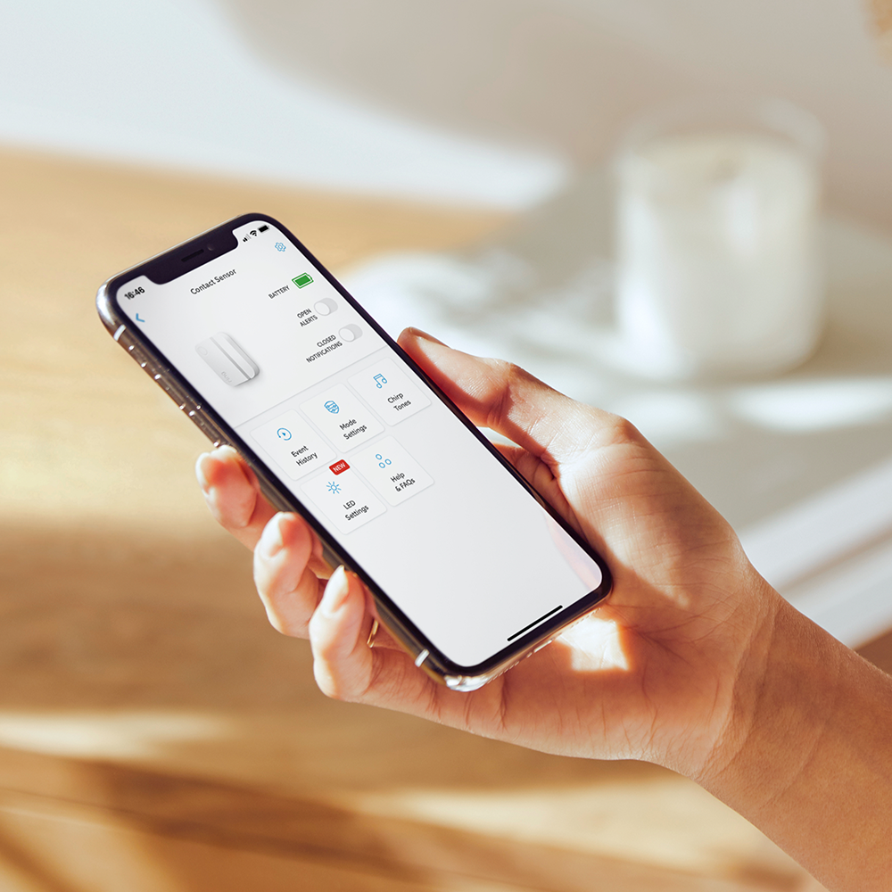 Hand holding a smartphone with an app showing a white door sensor. The phone displays the battery level.