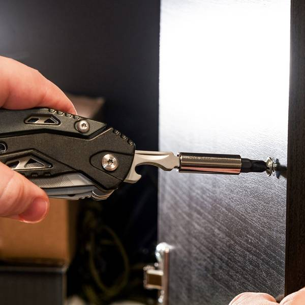 Kéz egy multitool-t használva, hogy becsavarjon egy csavart sötét fába. Az eszköz fekete és ezüst.