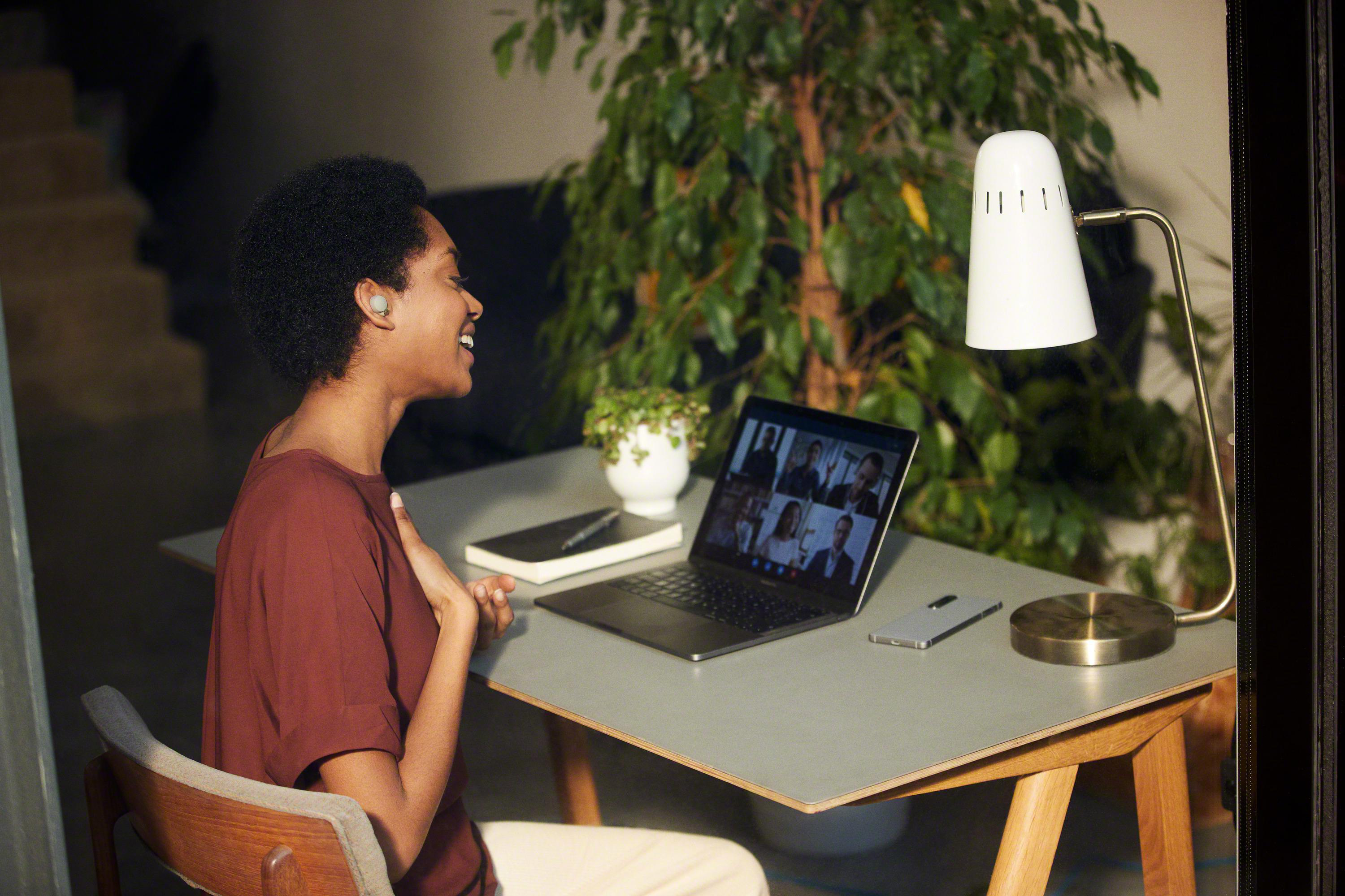 Eine Frau in einem Videoanruf lächelt. Ein Laptop, eine Lampe und eine Pflanze stehen auf dem Tisch.