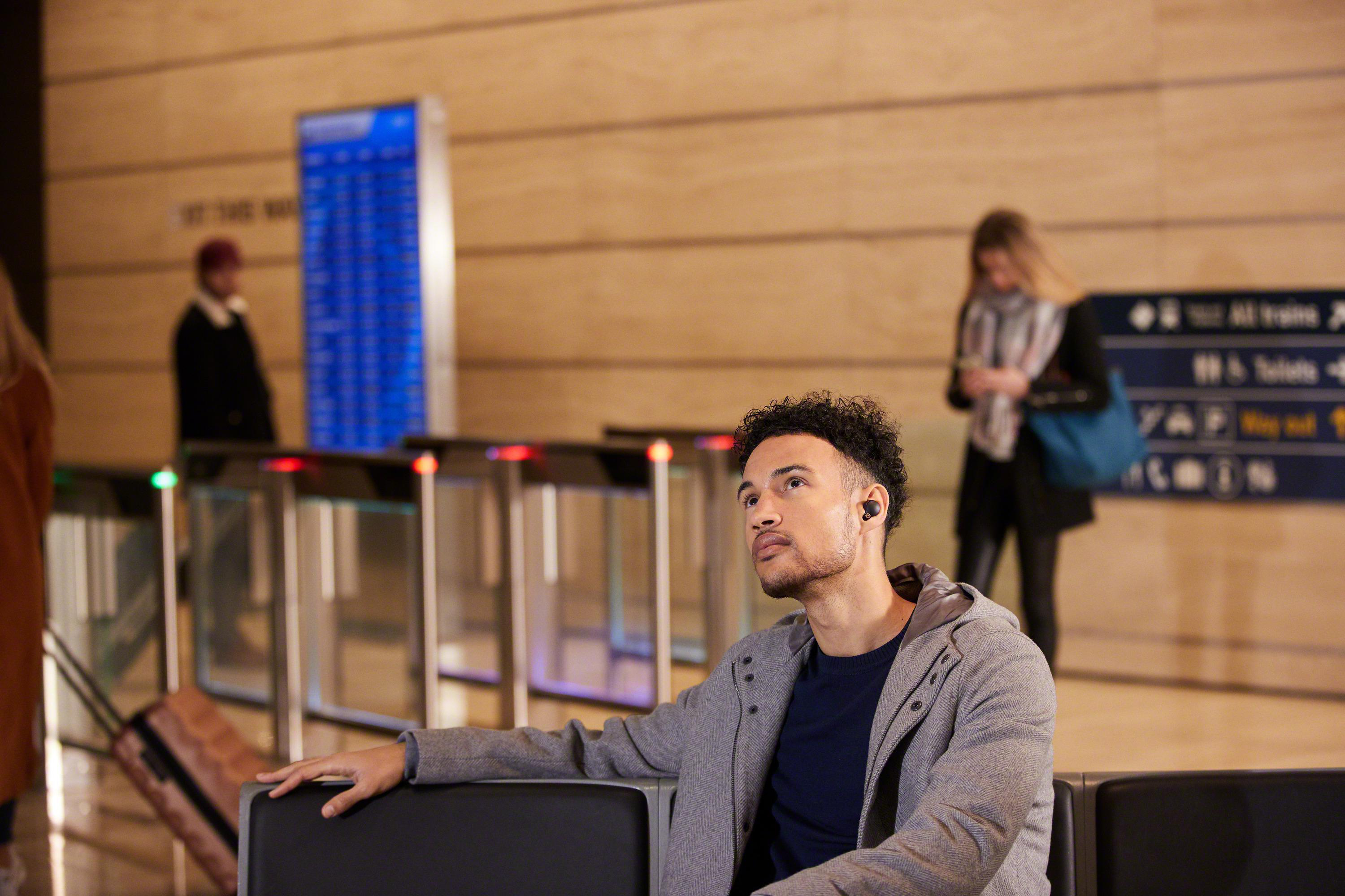 Mann in grauer Jacke mit Ohrhörern schaut in einem Flughafen nach oben. Leute und Schilder im Hintergrund.