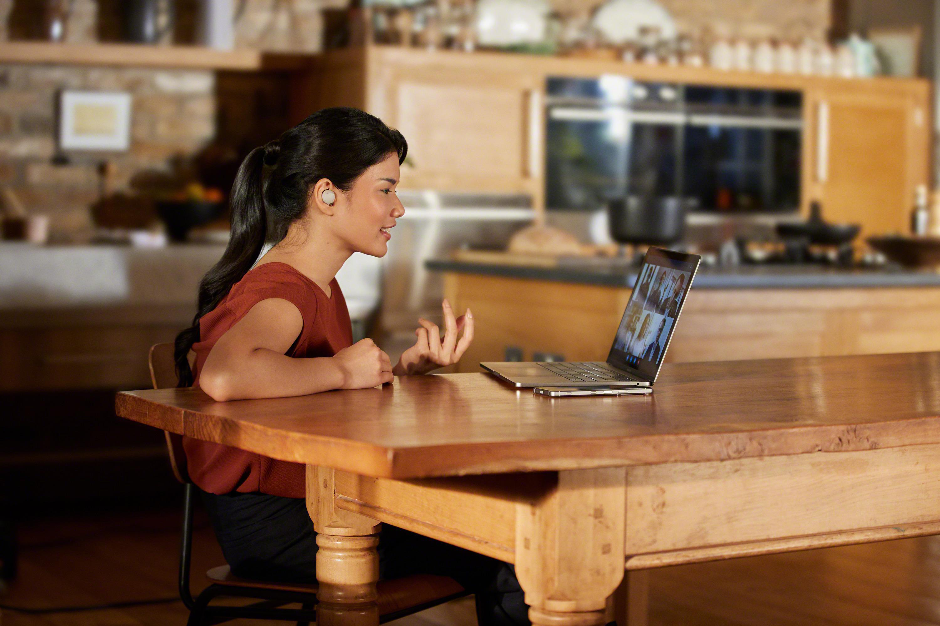 Frau im Headset bei Videokonferenz am Laptop, Holztisch, Küche im Hintergrund.