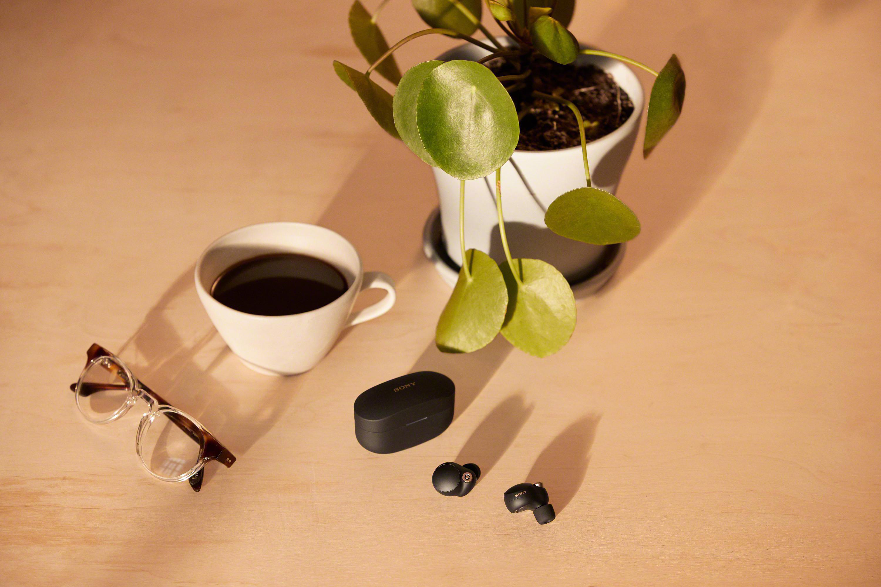 Eine Tasse Kaffee, Brille, Ohrhörer und eine Pflanze auf einer Holzoberfläche.